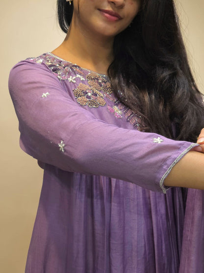 A woman wearing a lavender purple long-sleeve kurta with intricate silver and gold embroidery around the neckline and small embroidered motifs on the sleeves, shown against a plain beige background.