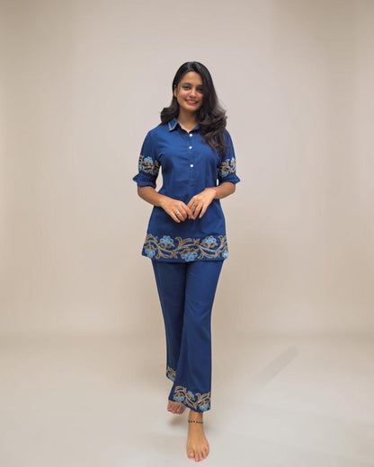 "Woman wearing a stylish blue embroidered co-ord set with palazzo pants, featuring floral detailing on the hem and sleeves, standing barefoot against a clean white background."
